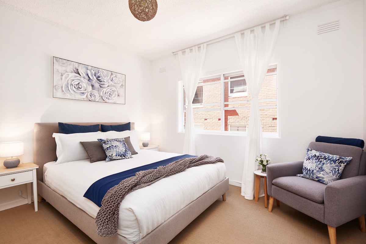 The bright bedroom features a spacious queen bed adorned with soft linens and decorative pillows. A plush armchair is positioned in the corner, accompanied by natural light filtering through sheer curtains. Two bedside tables with lamps provide an inviting atmosphere.