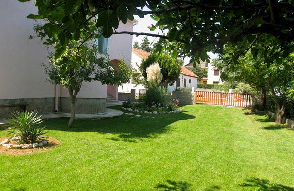 A green, well-maintained lawn is visible, with a variety of shrubs and small trees creating a soft border around the garden. A patio area is situated next to the house, and a wooden fence encloses the yard, providing a sense of privacy.