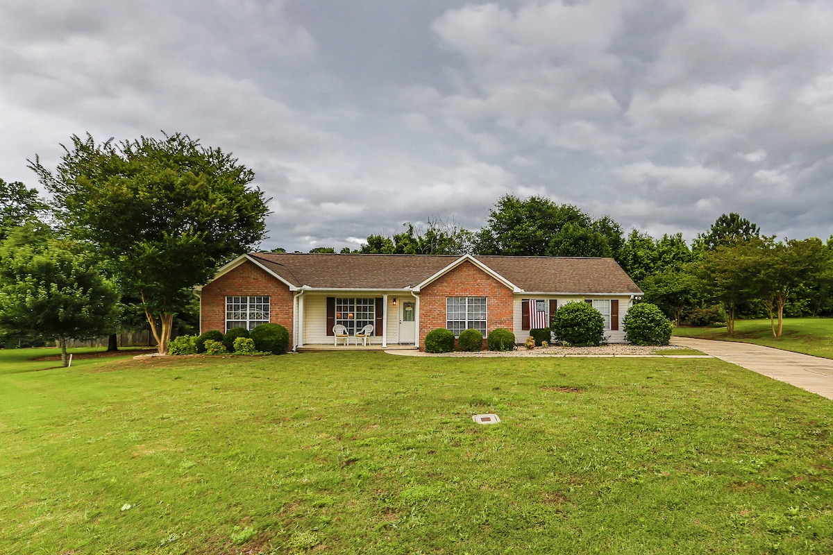 A well-maintained single-story home is set in a lush green landscape. The structure features a combination of brick and siding, complemented by neatly trimmed shrubs and a driveway leading to the entrance. Cloudy skies create a calm ambiance over the property.