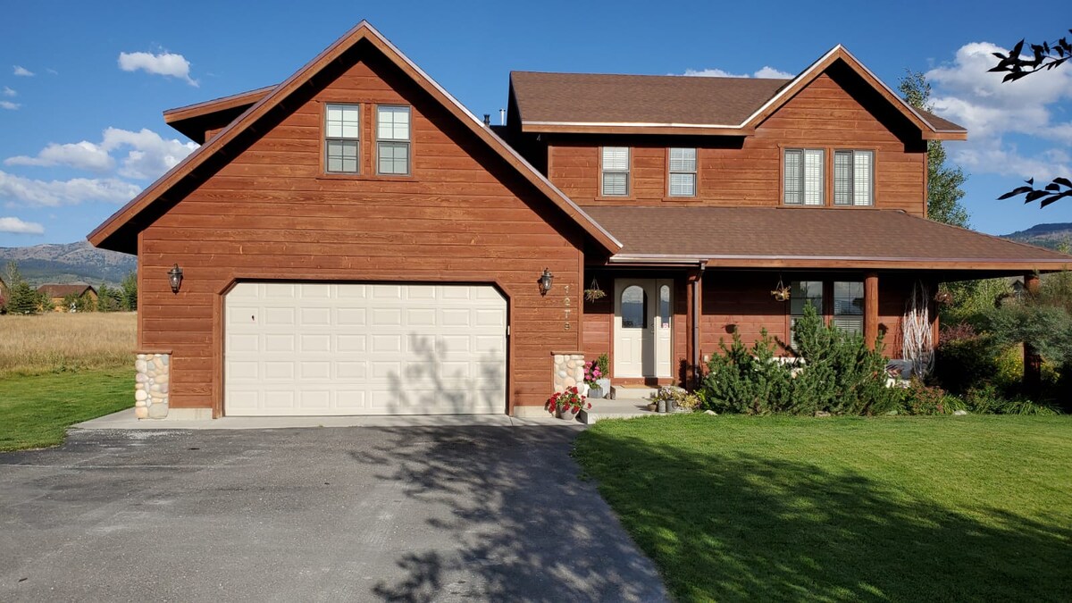 A spacious, two-story cabin features a warm wooden exterior with large windows. A front porch offers seating and flower arrangements, while an attached garage is visible. The surrounding landscape includes well-maintained grassy areas and gently rolling hills under a clear blue sky.
