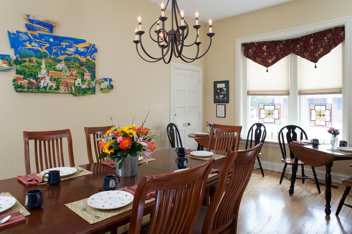 The dining room features a large wooden table surrounded by several chairs, set with plates and mugs. A vibrant floral arrangement sits in the center, while a stylish chandelier hangs above. Colorful window treatments complement decorative wall art, adding character to the space.