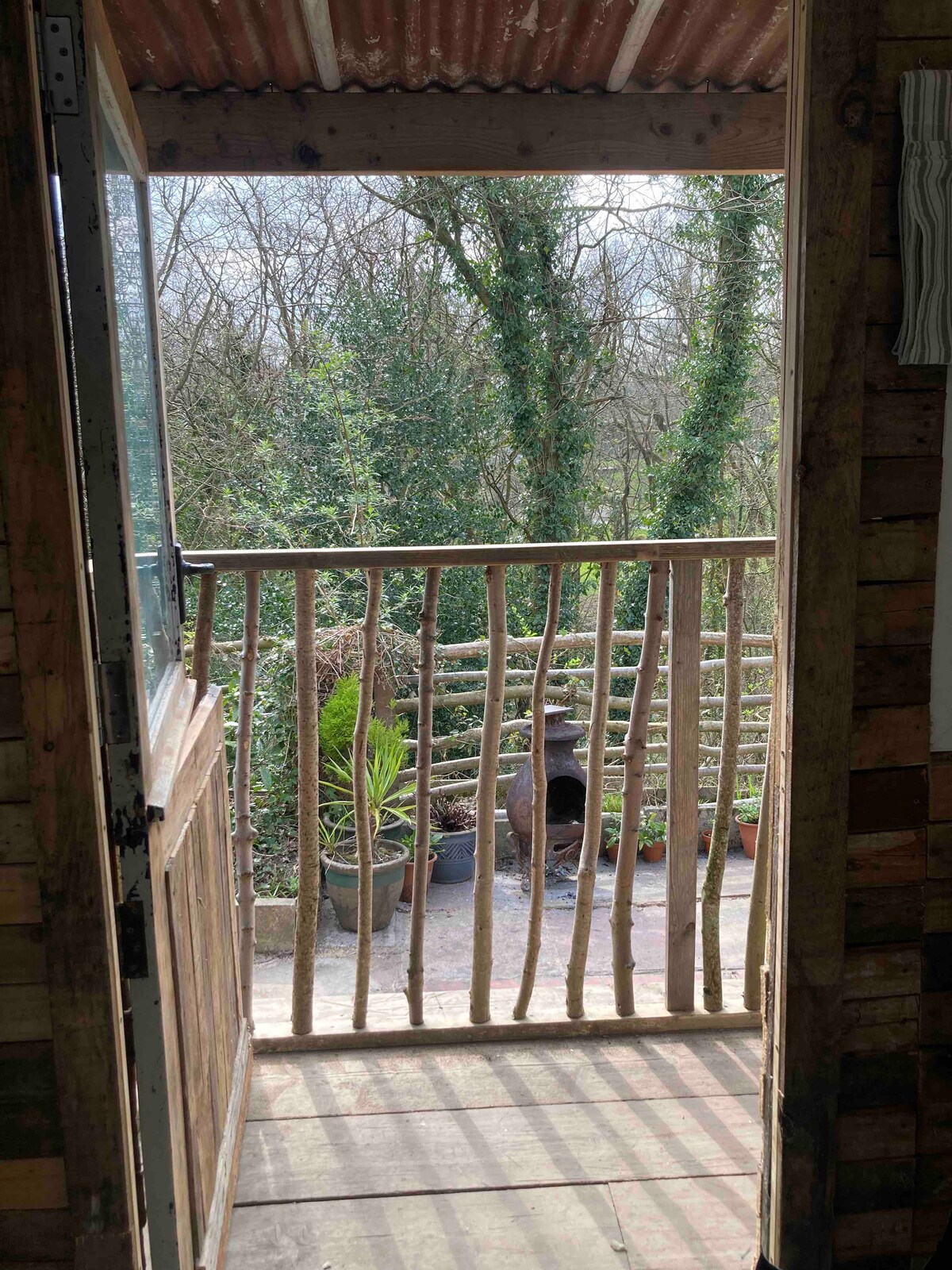 A wooden porch area features a rustic railing, opening to a natural landscape filled with greenery. Potted plants can be seen nearby, along with a fire pit positioned on the ground, creating an inviting outdoor space for relaxation.