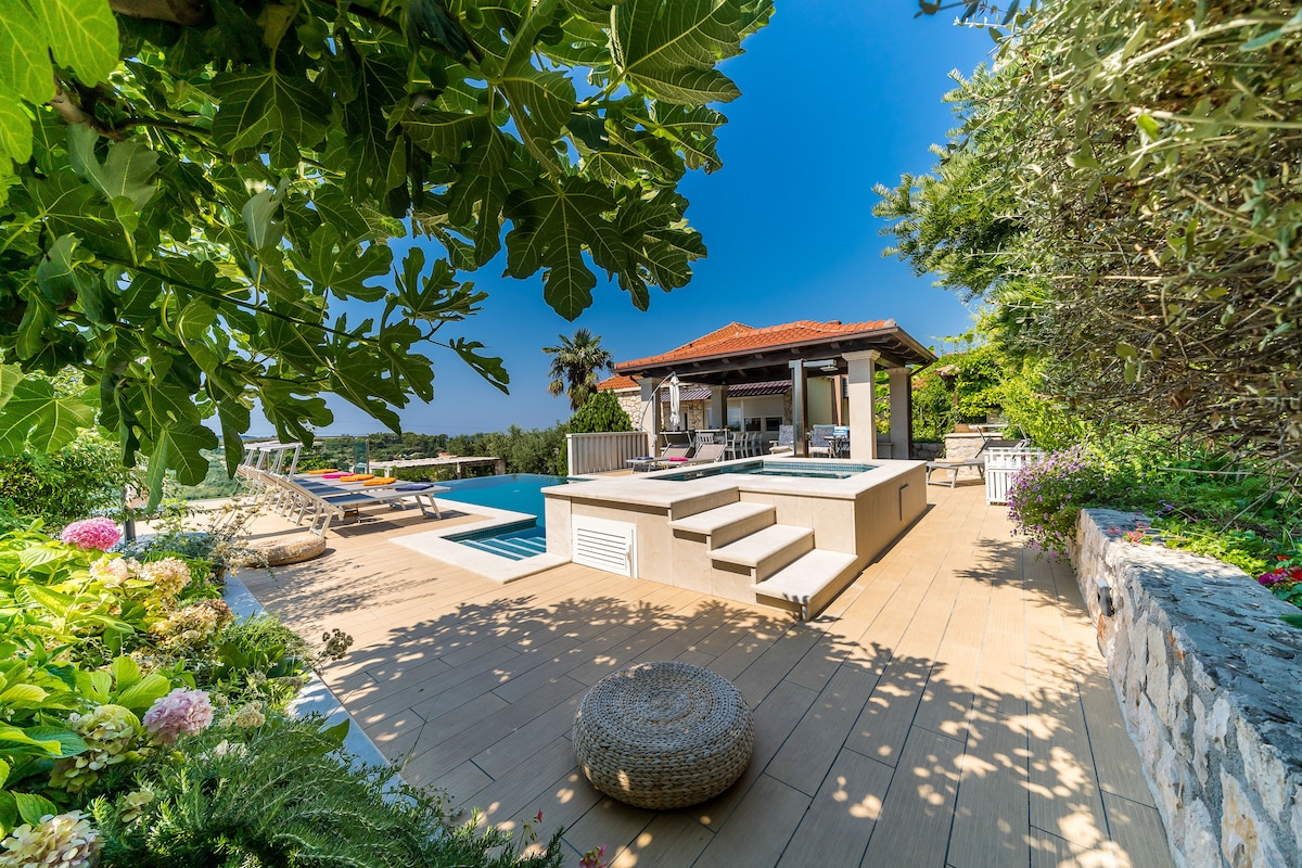 The outdoor area features a spacious pool surrounded by a well-maintained garden. A covered seating area is visible to the side, with a terrace made of wooden decking. Sun loungers line the poolside, providing a relaxing space for guests. A clear blue sky completes the serene setting.