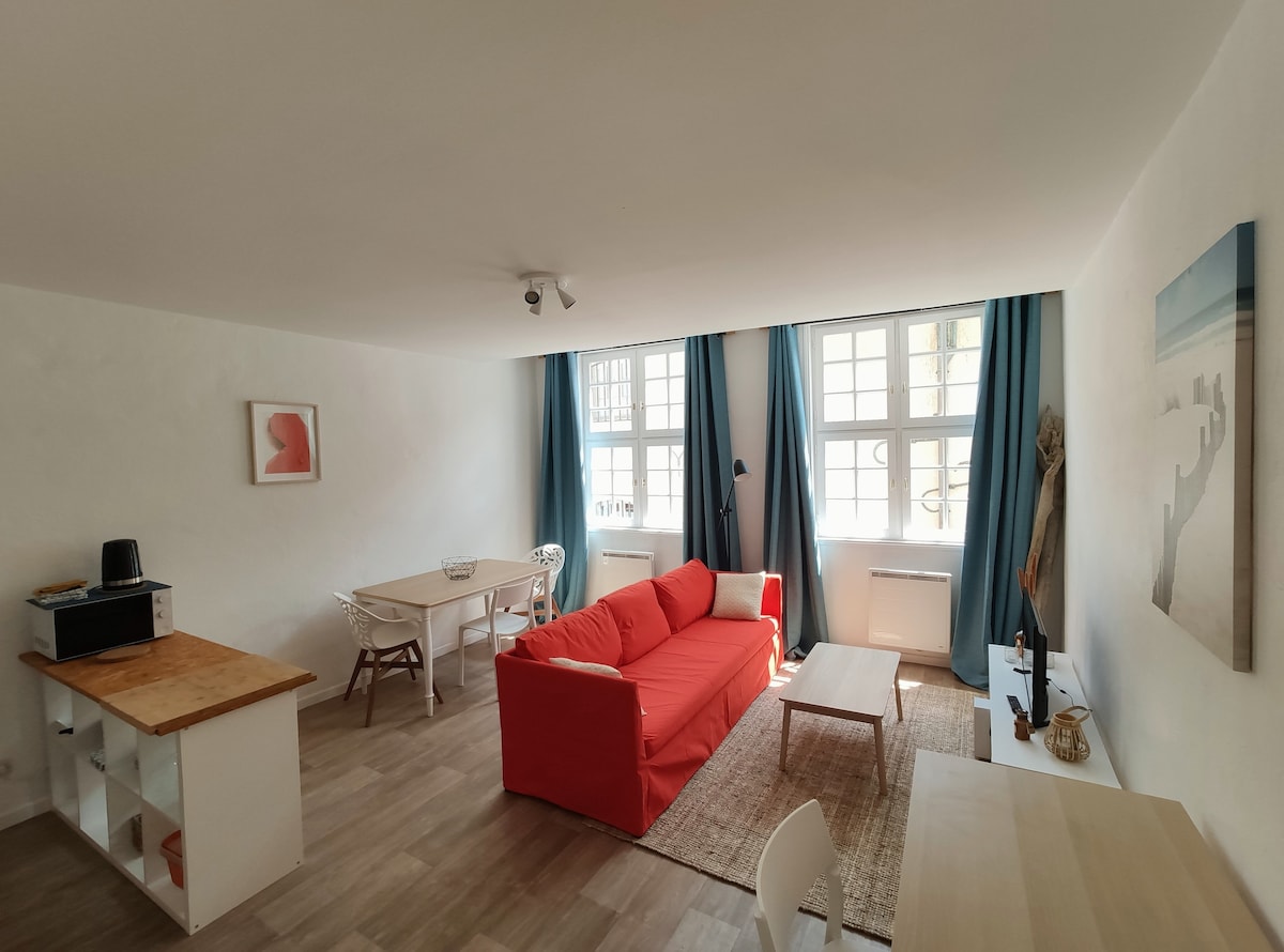 A spacious living area features a bright red sofa positioned in front of large windows, which let in ample natural light. A neutral rug is placed on the light wooden floor, with a dining table and chairs visible in the background, along with a small kitchenette.