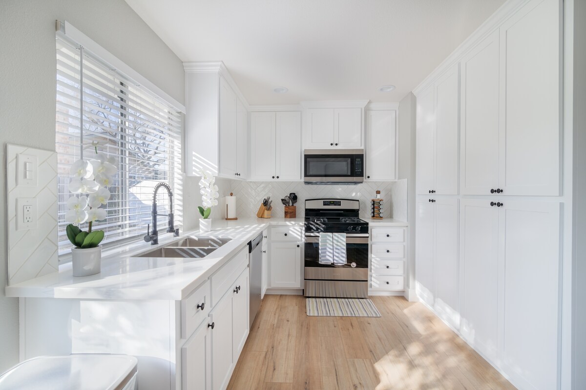 The bright kitchen features white cabinetry and a stainless steel stove. Natural light streams through the window, illuminating the countertop and modern sink. Open shelving displays decorative items, while a light-colored area rug adds warmth to the space.