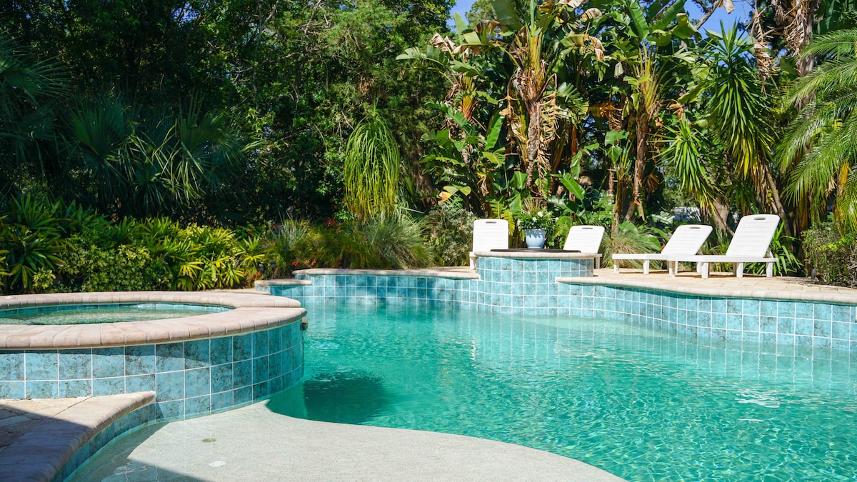 A serene saltwater pool glistens in the sunlight, surrounded by lush green foliage and tropical plants. A relaxing spa is positioned nearby, with two white lounge chairs set on the pool deck, inviting peaceful enjoyment of the outdoor space.