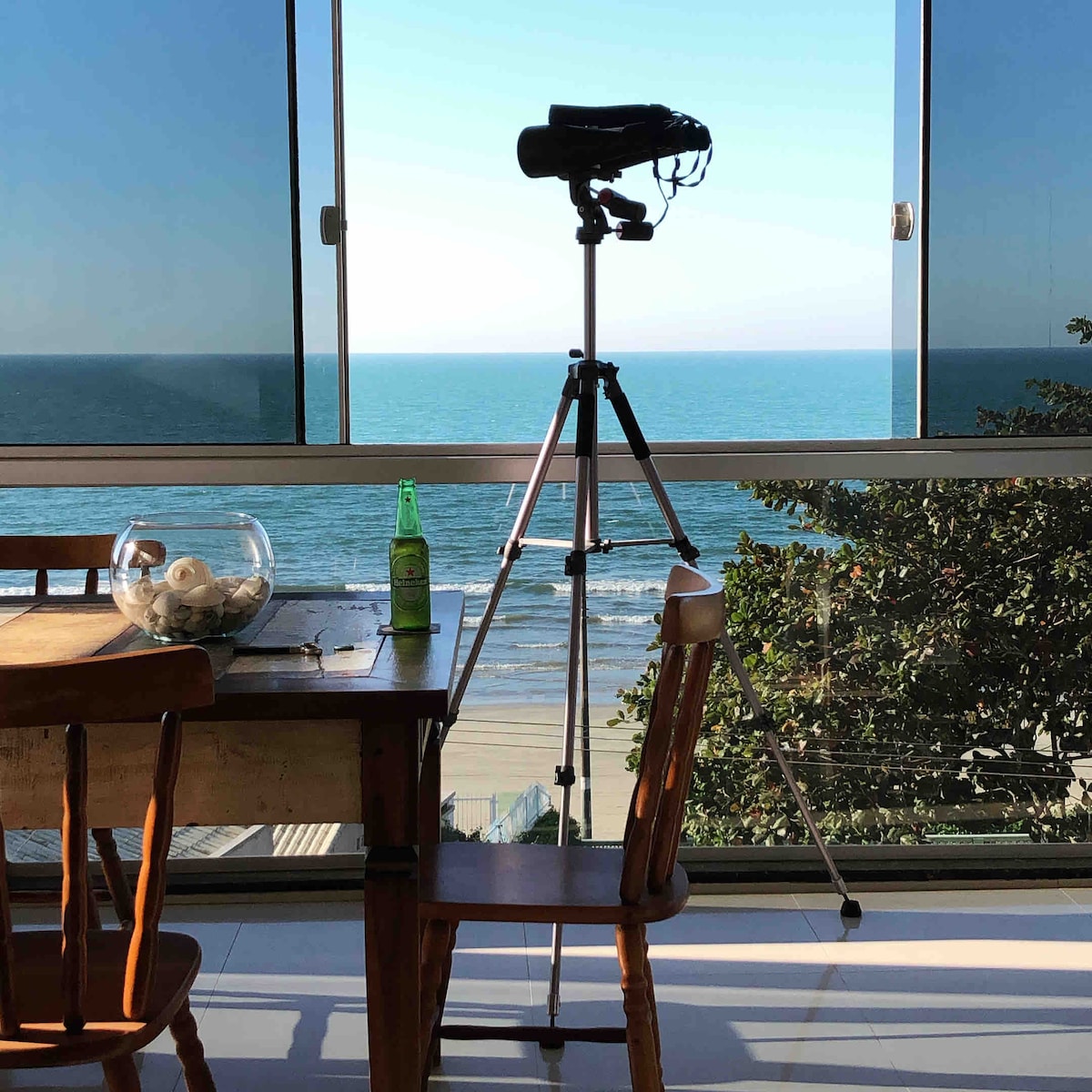 A dining area features a wooden table and a chair, framed by large glass windows that provide uninterrupted views of the beach and ocean. A telescope is positioned on a tripod, and a bottle sits on the table, enhancing the coastal ambiance.