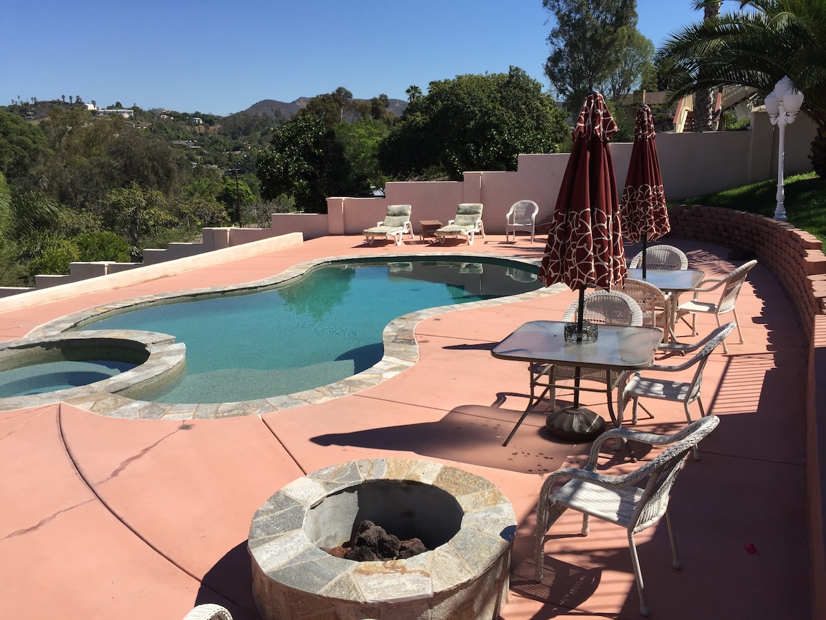 A spacious outdoor area features a solar-heated pool surrounded by a large patio. Several lounge chairs are positioned beside the pool, with two shaded tables nearby. A fire pit is visible, complementing the serene landscape of trees and hills in the background.