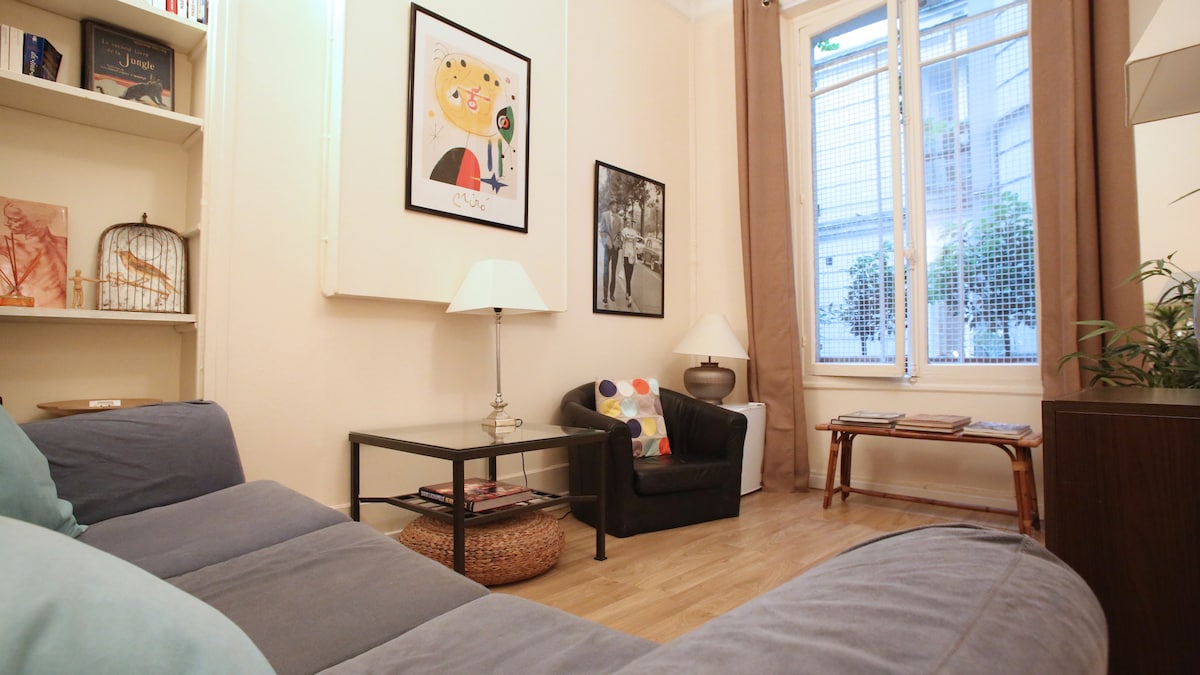 A cozy living space is shown, featuring a comfortable sofa with soft cushions and a glass coffee table. A lamp provides gentle lighting, while artwork adorns the walls. Natural light enters through a window with a view of the courtyard, and a small plant adds a touch of greenery.