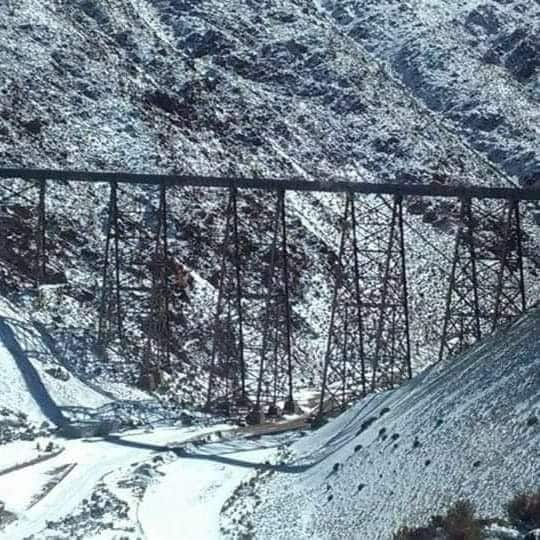 A large, multi-span bridge is visible, constructed of dark metal, crossing a snowy landscape. The surrounding mountains are covered in snow, highlighting the natural beauty of the area. A road can be seen below, winding through the terrain.