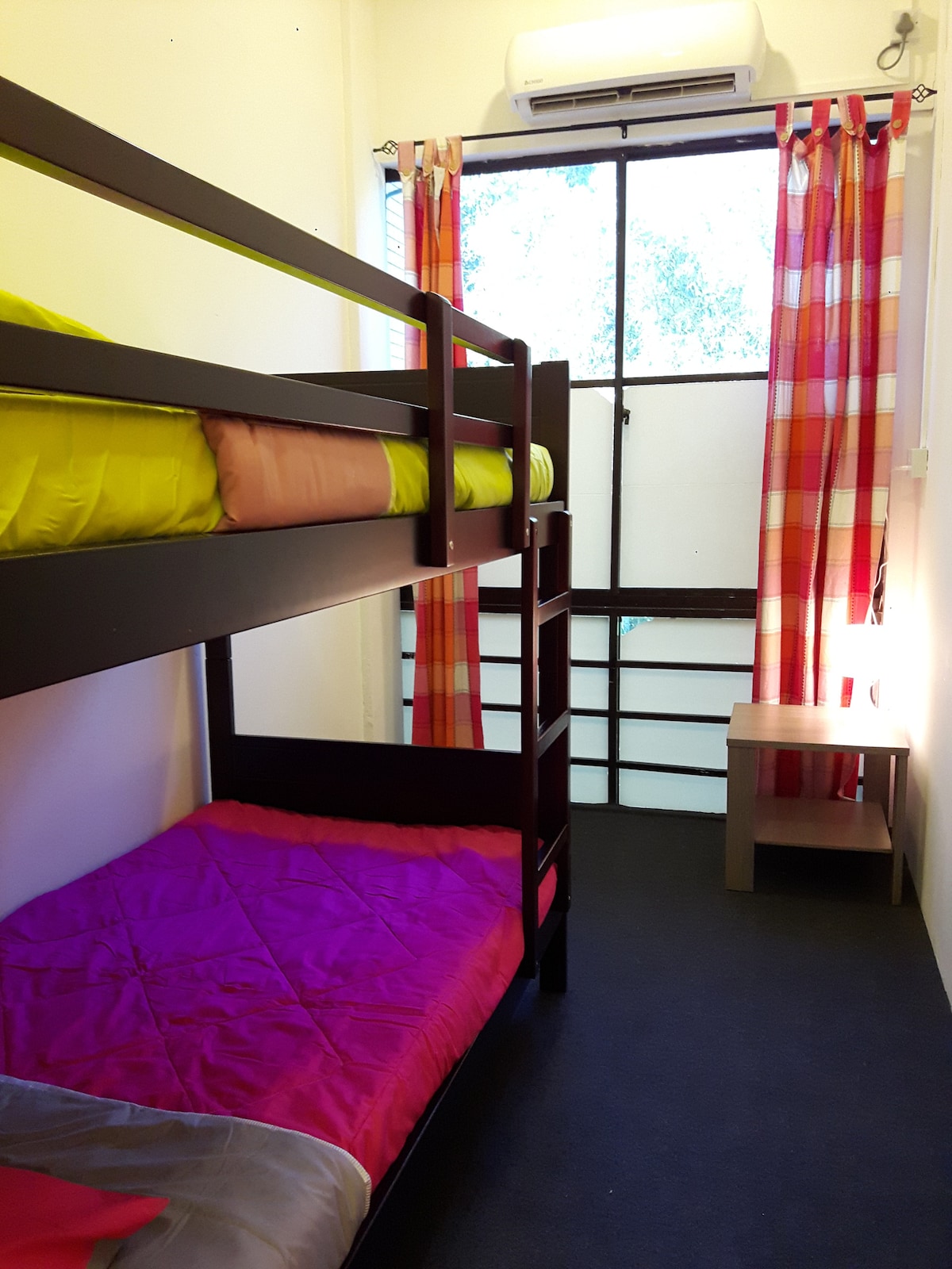 A bunk bed is positioned against the wall, featuring vibrant yellow and pink bedding. Colorful plaid curtains frame the window, allowing natural light to enter. A small bedside table is placed beside the lower bunk, enhancing the cozy space.