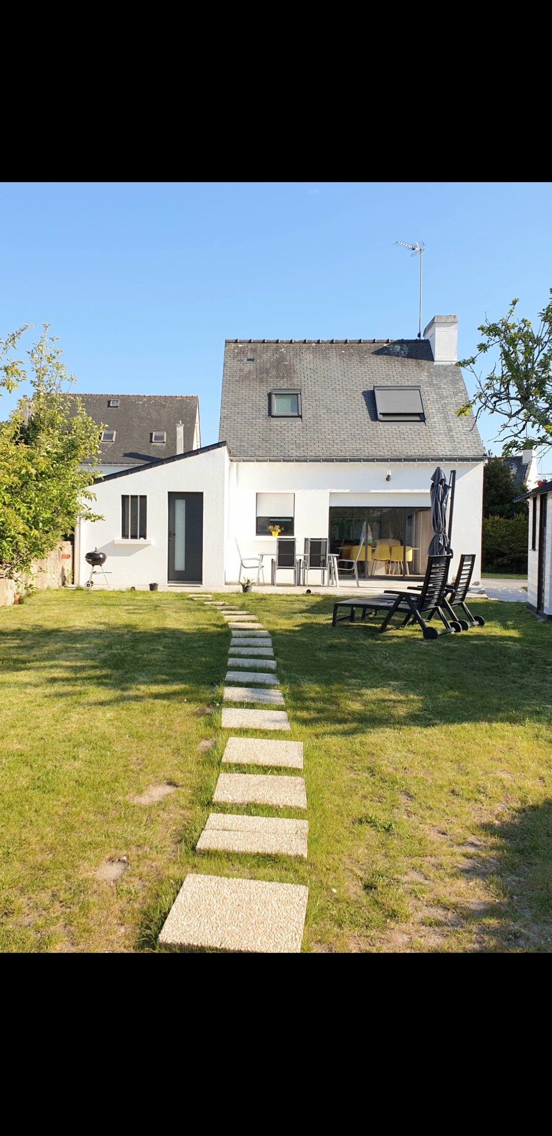 The exterior of a two-story house is framed by a grassy lawn, with a walkway of square stone tiles leading to the entrance. Outdoor seating is displayed on a small terrace, accompanied by a lounge chair. A clear sky complements the bright and open setting.