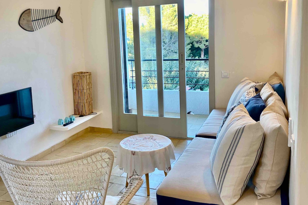 A bright living area features a spacious sofa adorned with neutral and blue-striped cushions. A small round table is set beside, while a flat-screen TV is mounted on the wall. Large glass doors lead to a balcony with a view of greenery.