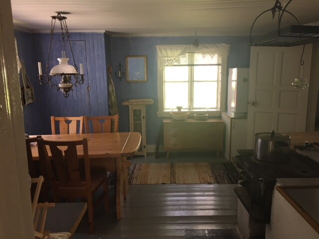 A rustic kitchen and dining area features wooden furniture, including a table with several chairs. A large window allows natural light to fill the space, highlighting the blue walls. A vintage stove and a gas fridge are visible, contributing to the homely atmosphere.