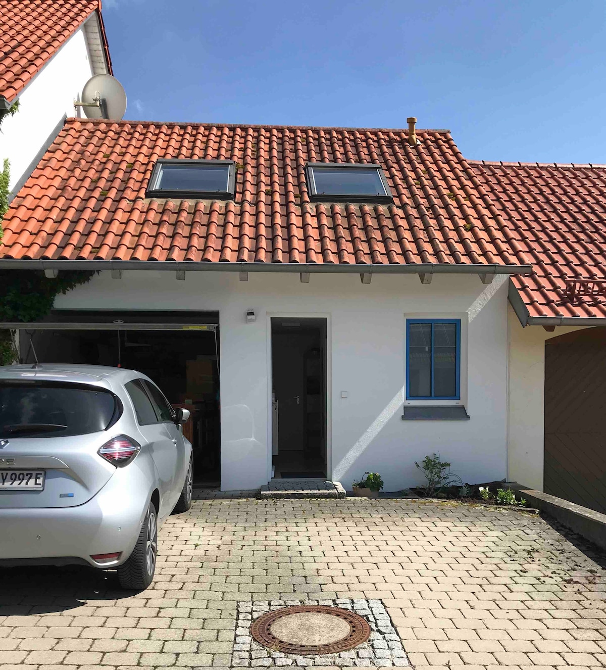 The exterior of a house is shown, featuring a sloped roof covered in red tiles. Two skylights are visible on the upper level. A white car is parked in front of the entrance, which has a gray stone pathway leading to the front door. A small garden area is present beside the entrance.