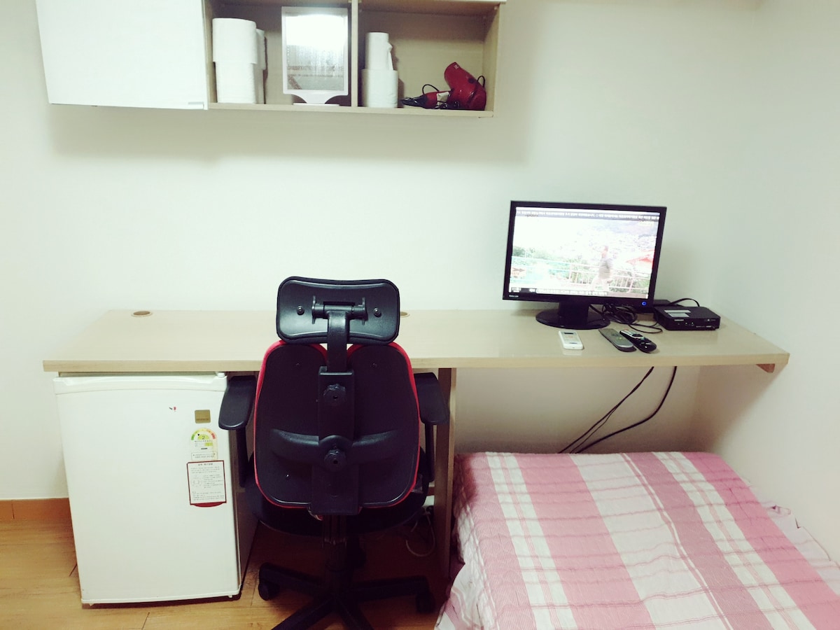 A functional workspace is arranged with a desk, a chair, and a monitor. A small refrigerator is positioned nearby, and a neatly made bed with a checkered blanket is visible. Above, a shelf holds essentials, including paper towels and other items.