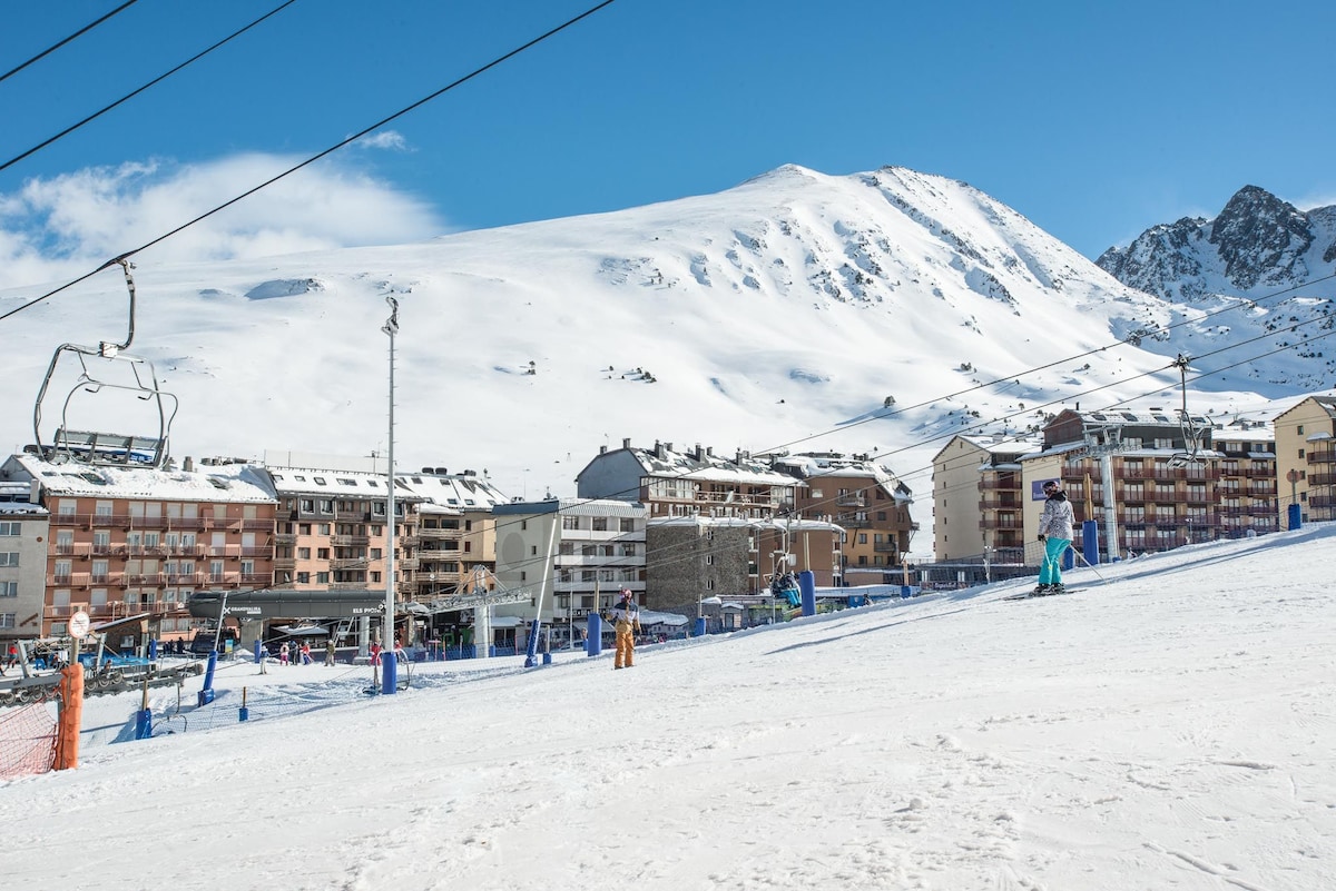 A snowy landscape is visible, showcasing ski slopes with a backdrop of majestic mountains. Nearby buildings, displaying a variety of architectural styles, are situated along the base of the slopes. Clear blue skies enhance the winter scene, creating a serene atmosphere for winter sports.