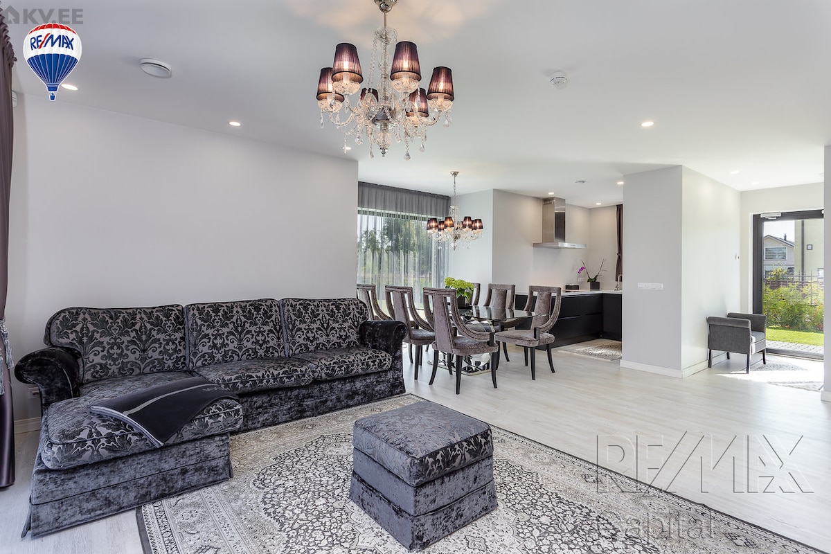 A spacious living area features a dark velvet sofa and an elegant chandelier. A dining table with six chairs is visible in the background, alongside a modern kitchen. Natural light enters through large windows, enhancing the open concept design.
