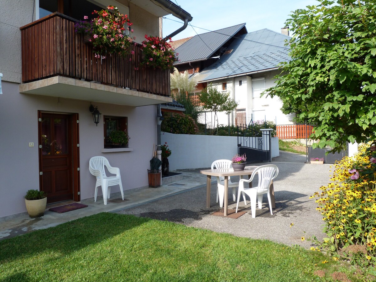 A tranquil outdoor area features a small wooden table surrounded by two white chairs on a grassy surface. An entrance door is visible, flanked by pots with vibrant flowers. A canopy of trees and shrubs provides a natural backdrop.