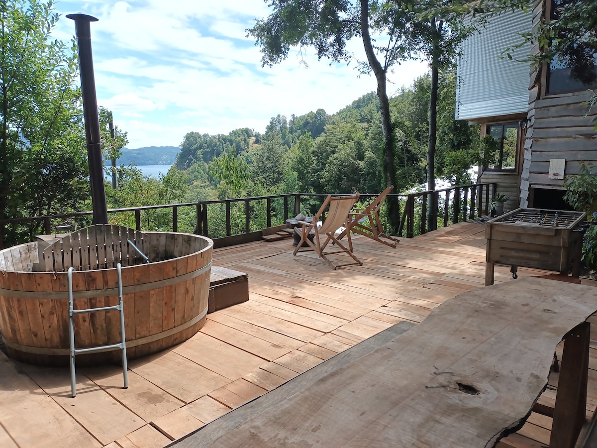 A spacious wooden deck features a hot tub surrounded by greenery, offering a view of the lake in the distance. Two lounge chairs are positioned for relaxation, and a rustic wooden table adds to the inviting outdoor space.