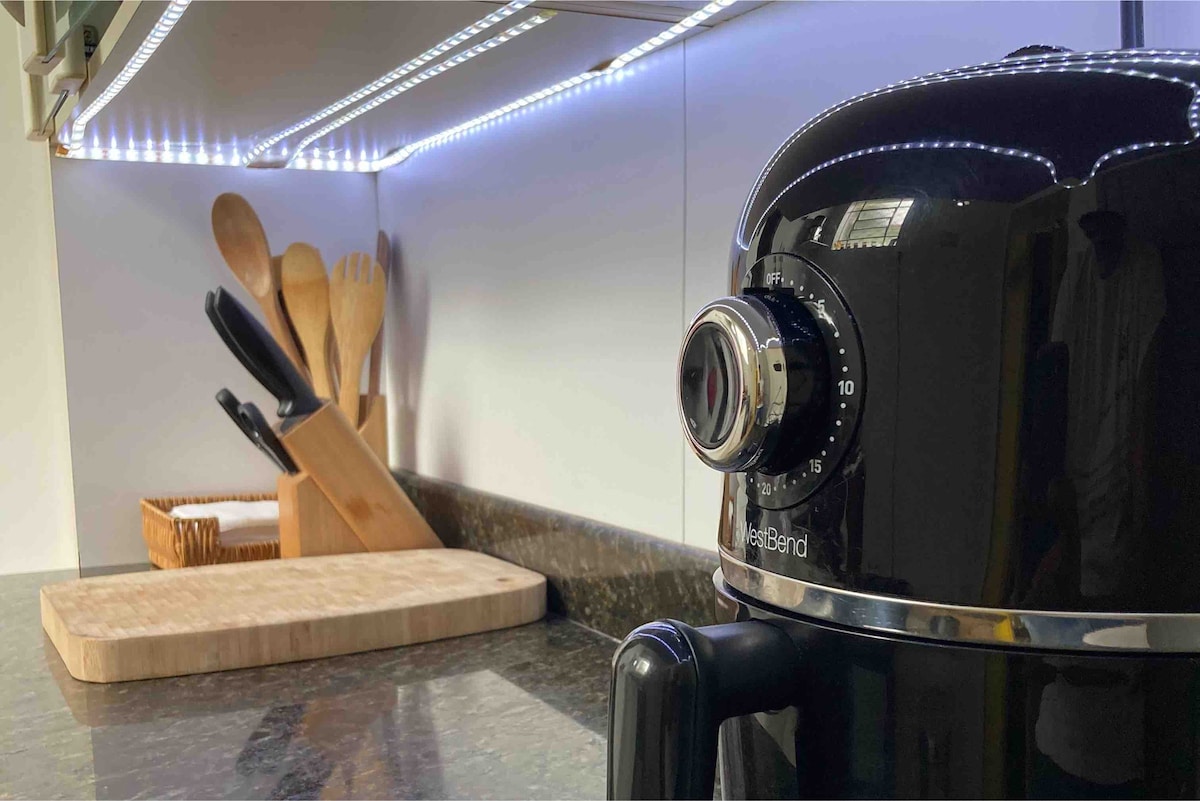 A sleek black air fryer is prominently displayed on a dark countertop, reflecting the modern kitchen's style. Behind it, utensils and knives are organized neatly on a wooden holder, with soft lighting enhancing the workspace.