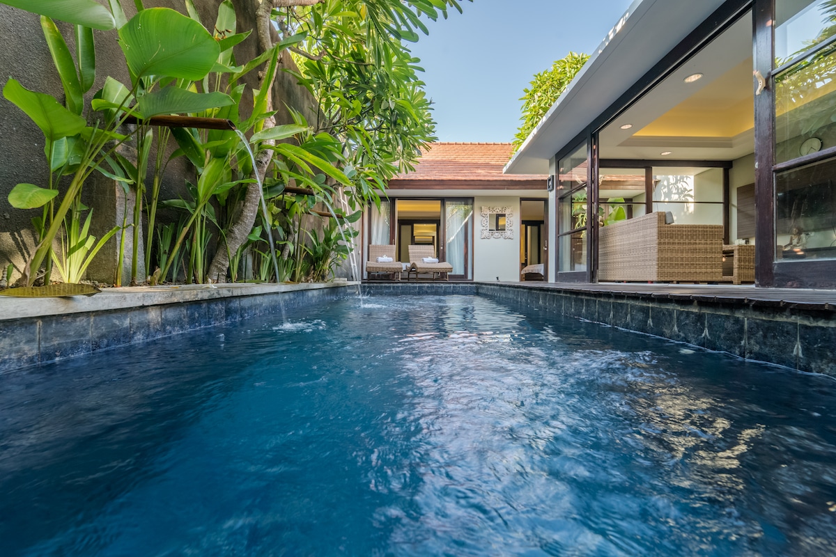 The image showcases a private pool with clear, inviting water bordered by lush tropical plants and a wooden deck. In the background, the open living space features large glass doors, promoting a seamless indoor-outdoor experience, with comfortable seating visible.