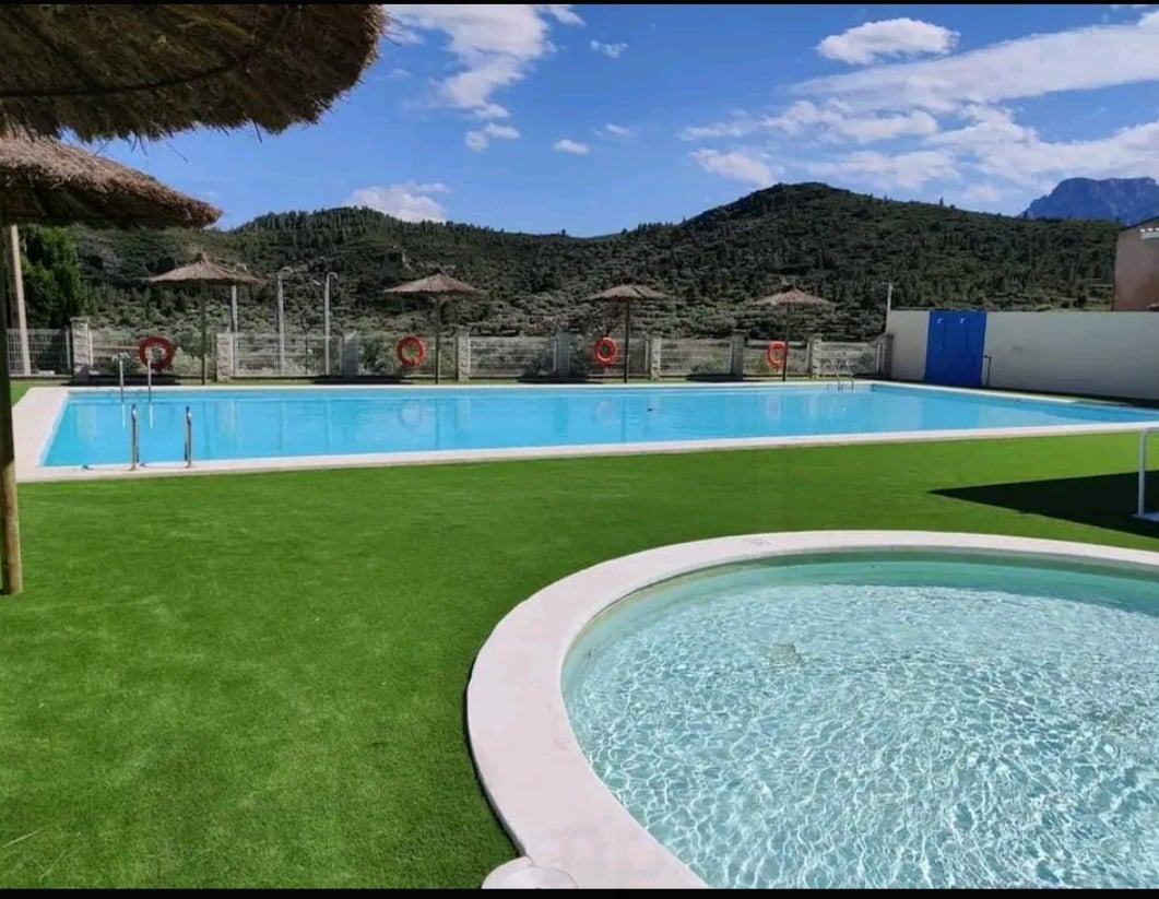 A large swimming pool with clear blue water is surrounded by green grass and lounge chairs. Shade is provided by thatched umbrellas. A smaller round spa pool is visible nearby, creating a relaxing atmosphere with beautiful mountain views in the background.