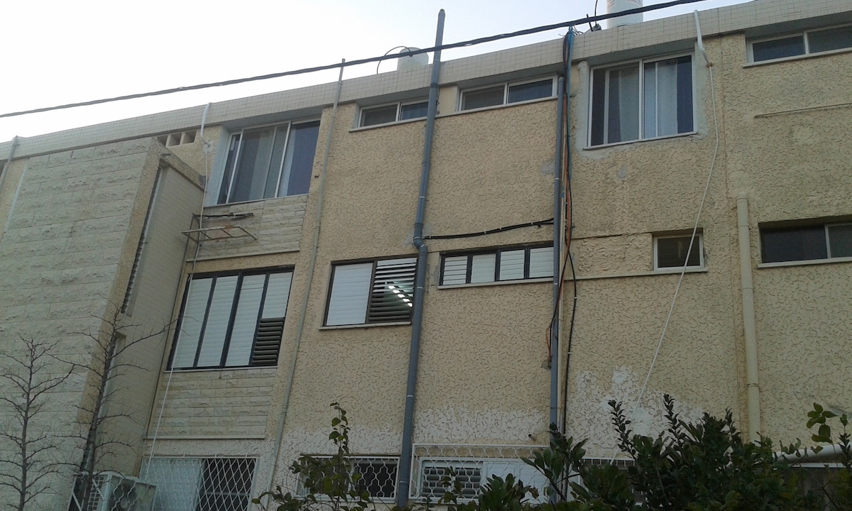 The exterior of a multi-story building is displayed, featuring a textured façade in a light color. Windows and balconies are visible, with some partially open. A small garden with shrubs is located at the base, and utility lines are affixed to the side of the structure.