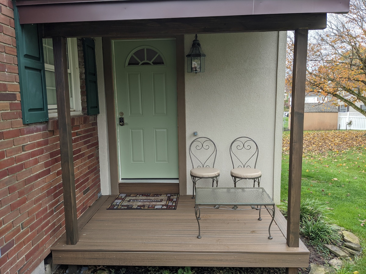 A welcoming porch features a light green front door framed by wooden posts. Two intricate wrought iron chairs sit beside a glass-topped table, creating a cozy spot for refreshments. Vibrant autumn leaves are visible in the background, enhancing the outdoor appeal.