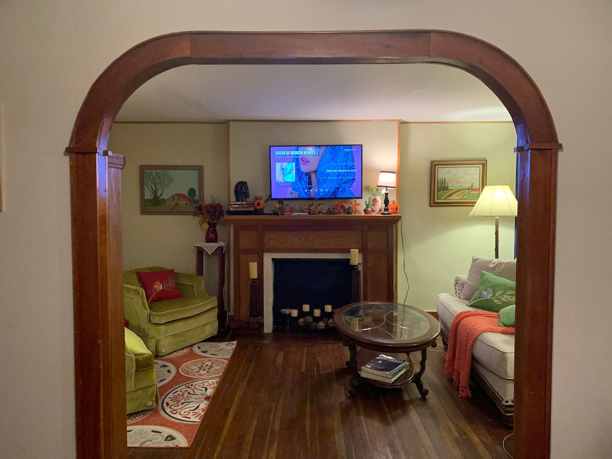 A welcoming living room is framed by an arched entryway, featuring a cozy couch adorned with colorful blankets and a green armchair. A round coffee table sits on a patterned rug. The fireplace is complemented by art pieces and soft lighting, creating a relaxed ambiance.