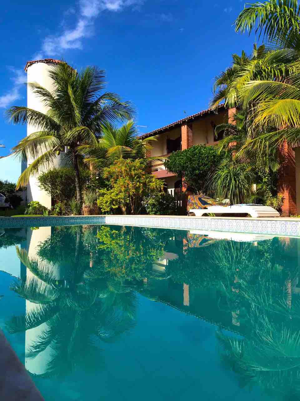 A tranquil swimming pool reflects the surrounding greenery and architecture, with palm trees and vibrant plants framing the area. The two-story building is visible in the background, contributing to the serene atmosphere. Clear blue skies enhance the peaceful setting.