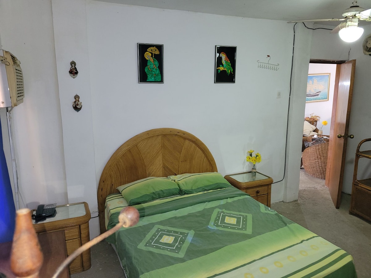 A simple bedroom is featured, showcasing a spacious bed with green bedding. Two paintings adorn the white walls above, while a bedside table sits on either side. An air conditioning unit is mounted on the wall, and a door leads to another room.