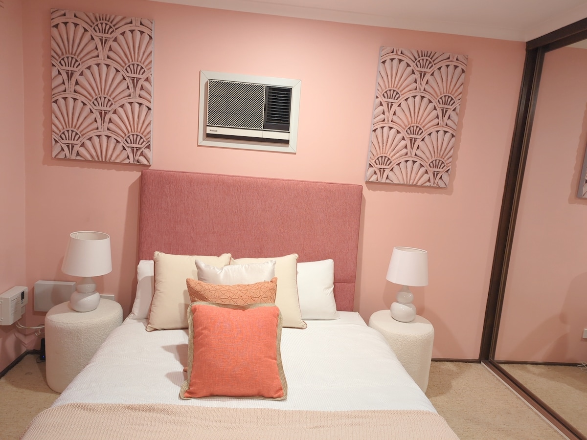 A calming bedroom ambiance is created by soft pink walls and decorative wall accents. The spacious bed is adorned with assorted throw pillows and a light blanket. Nightstands with lamps flank the bed, and a mirrored wardrobe reflects the cozy setting.