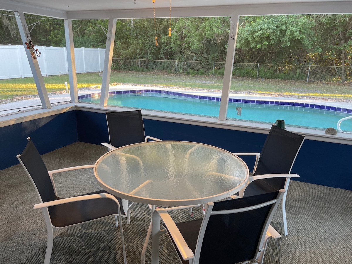 A screened lanai features a glass-top dining table surrounded by four black chairs. The space offers a clear view of a swimming pool and the lush green backyard beyond, with natural lighting coming through the large windows.