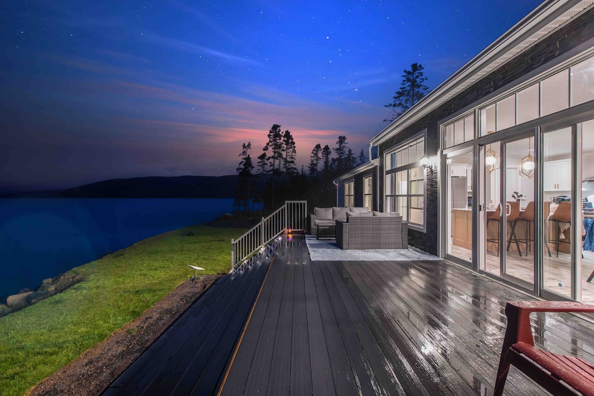 An outdoor deck extends toward the water, showcasing comfortable seating and scenic views of the ocean and mountains under a twilight sky. Soft lighting from the interior illuminates the space, enhancing the serene atmosphere.