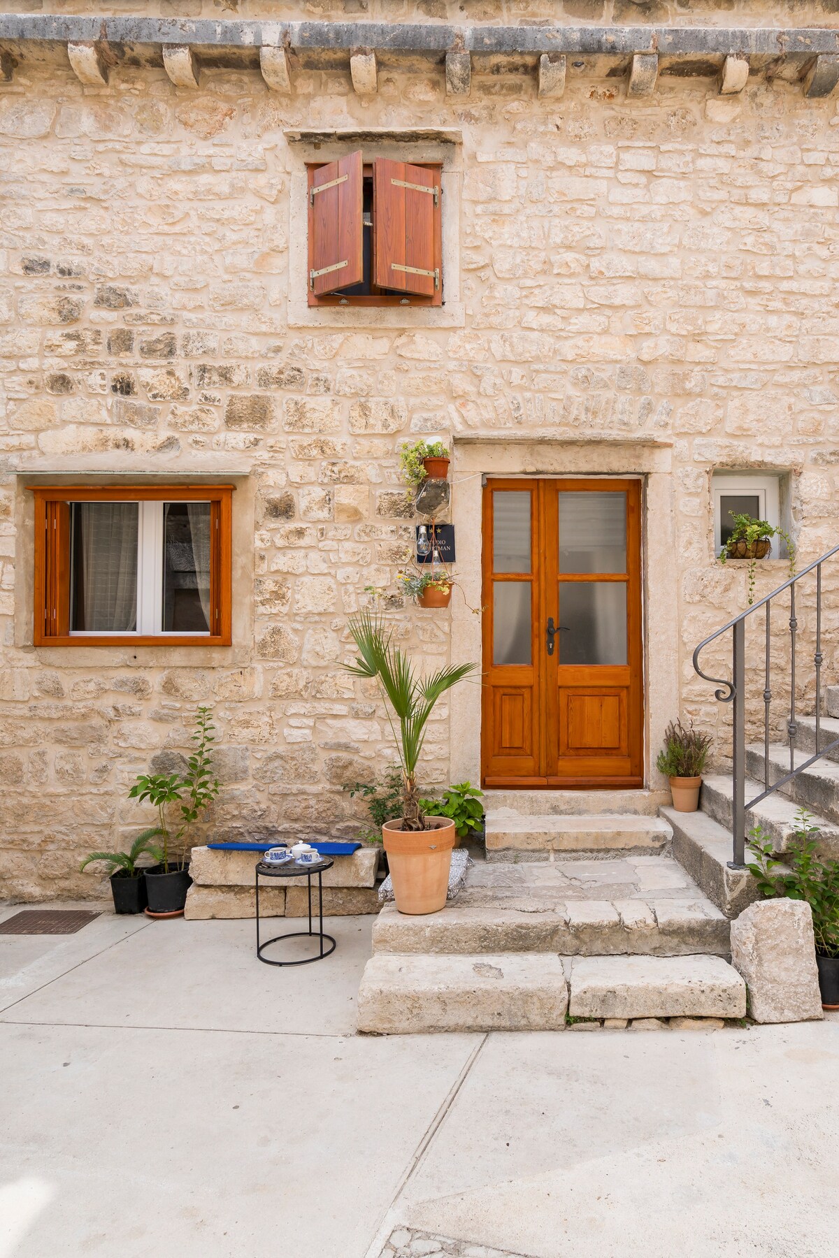 The exterior features a stone facade with a warm wooden door surrounded by planters. A small table is placed on the patio steps, accompanied by a potted plant. The second window has wooden shutters, enhancing the building's rustic charm.