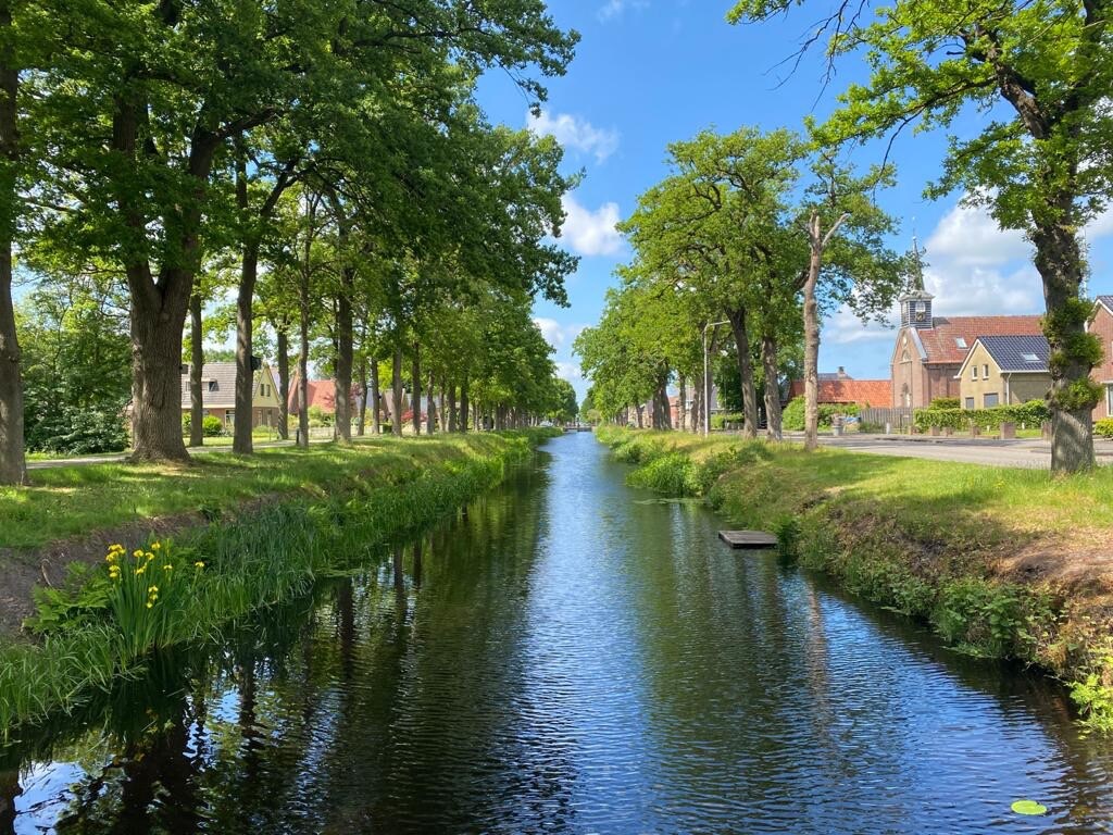 A serene view of a canal is framed by lush trees lining both sides. The clear water reflects the blue sky and cotton-like clouds. In the distance, charming buildings can be seen, enhancing the natural beauty of the area.
