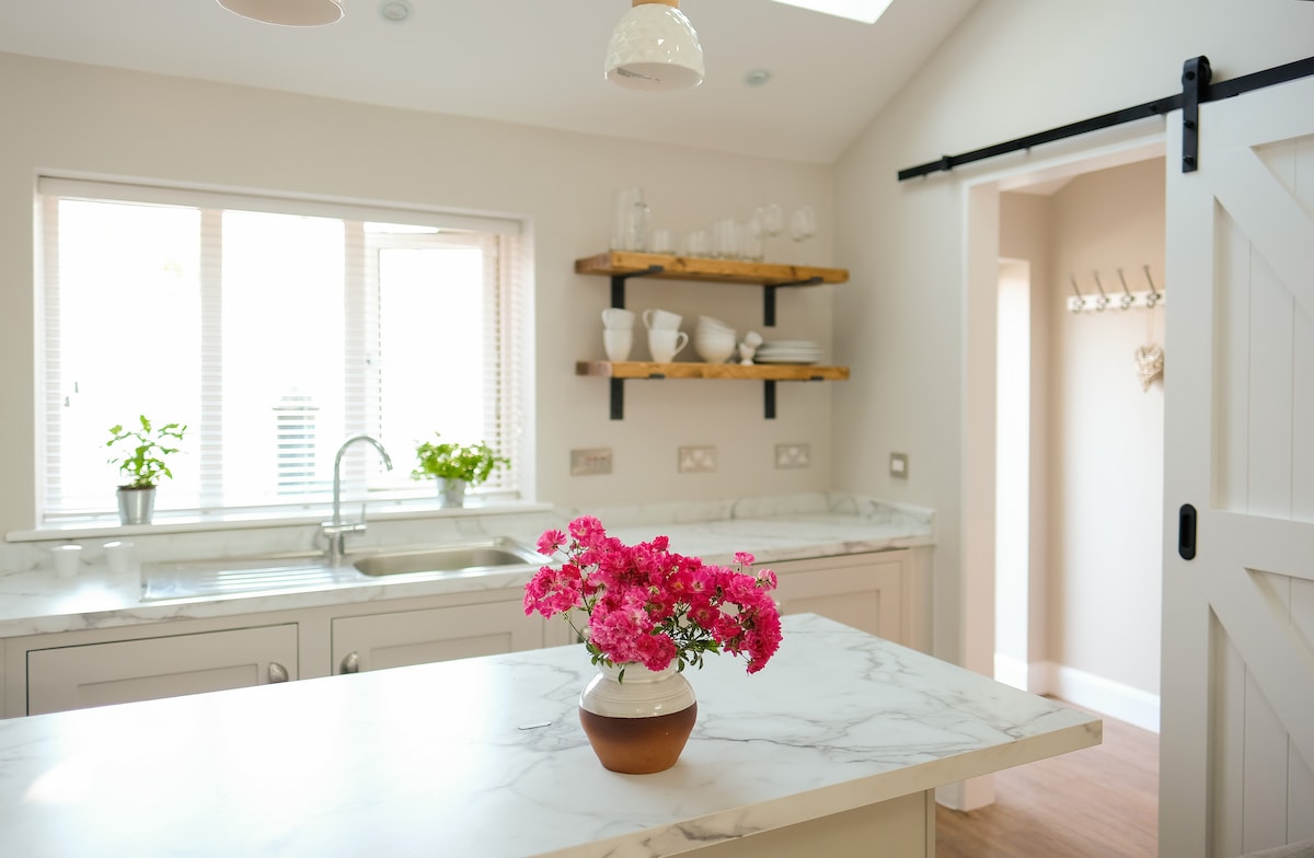 A bright kitchen features a marble island with a floral arrangement in a vase. Natural light filters through the window, illuminating the contemporary cabinetry and wooden shelves lined with dishware. A sink is positioned beneath the window, providing an inviting workspace.