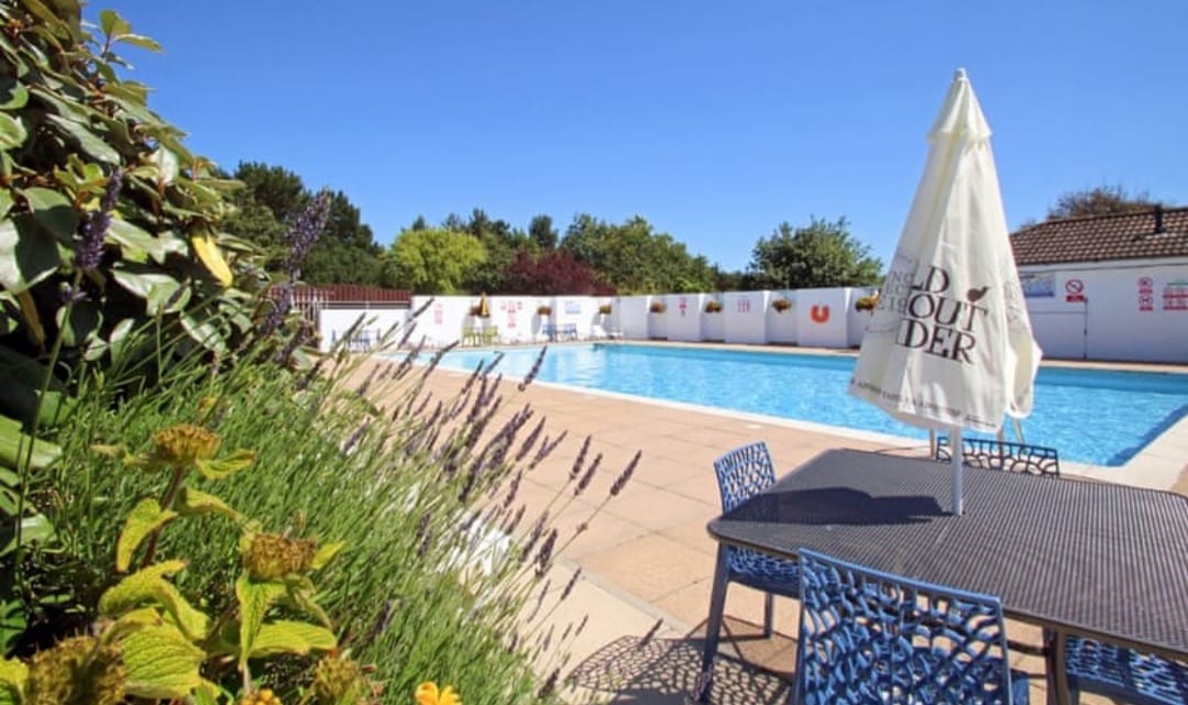 An outdoor swimming pool is surrounded by a sunlit patio, featuring deck chairs and a table with vibrant blue chairs. Lush greenery and flowers are visible in the foreground, adding natural beauty to the area. A bright sky completes the inviting atmosphere.