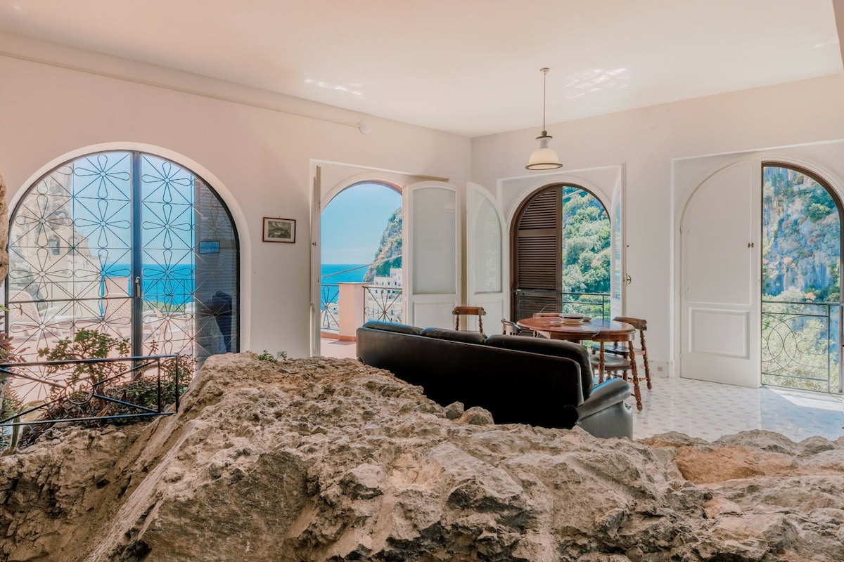 The living area features arched doorways opening up to stunning sea views, highlighting the connection between indoor and outdoor spaces. A dark sofa faces a wooden dining table, while natural rock elements create a unique focal point. Soft light filters in through the open doors.