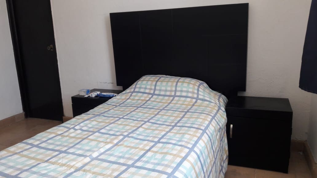 A neatly arranged sleeping area features a bed with a patterned blanket. A dark headboard complements the room's color scheme. A nightstand is positioned beside the bed, providing surface space. Light beige tiles cover the floor, contributing to a clean and simple atmosphere.