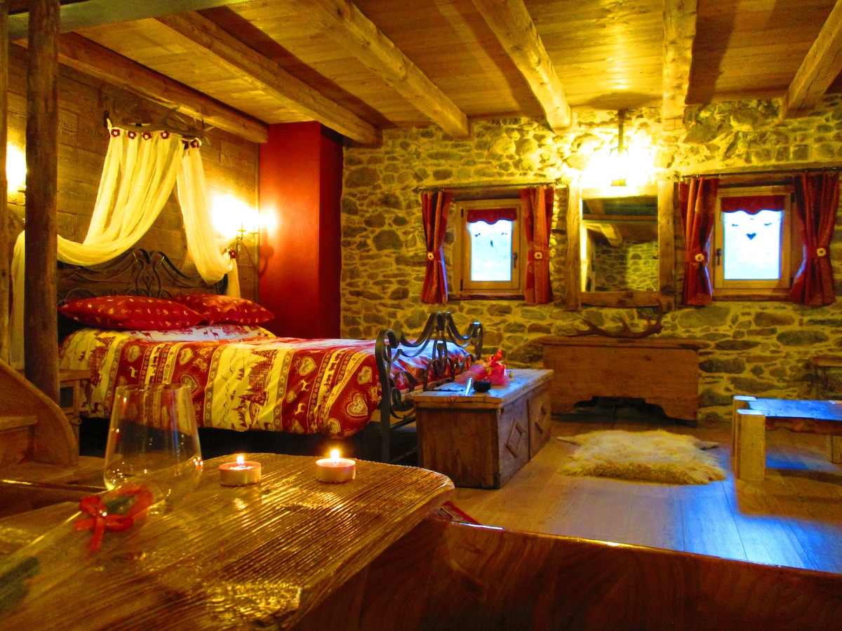 The spacious bedroom features an intricately designed metal bed adorned with rich red bedding. Stone walls contrast with warm wooden beams overhead. Soft lighting illuminates the area, highlighting a wooden dresser and a cozy seating space, inviting relaxation and comfort.