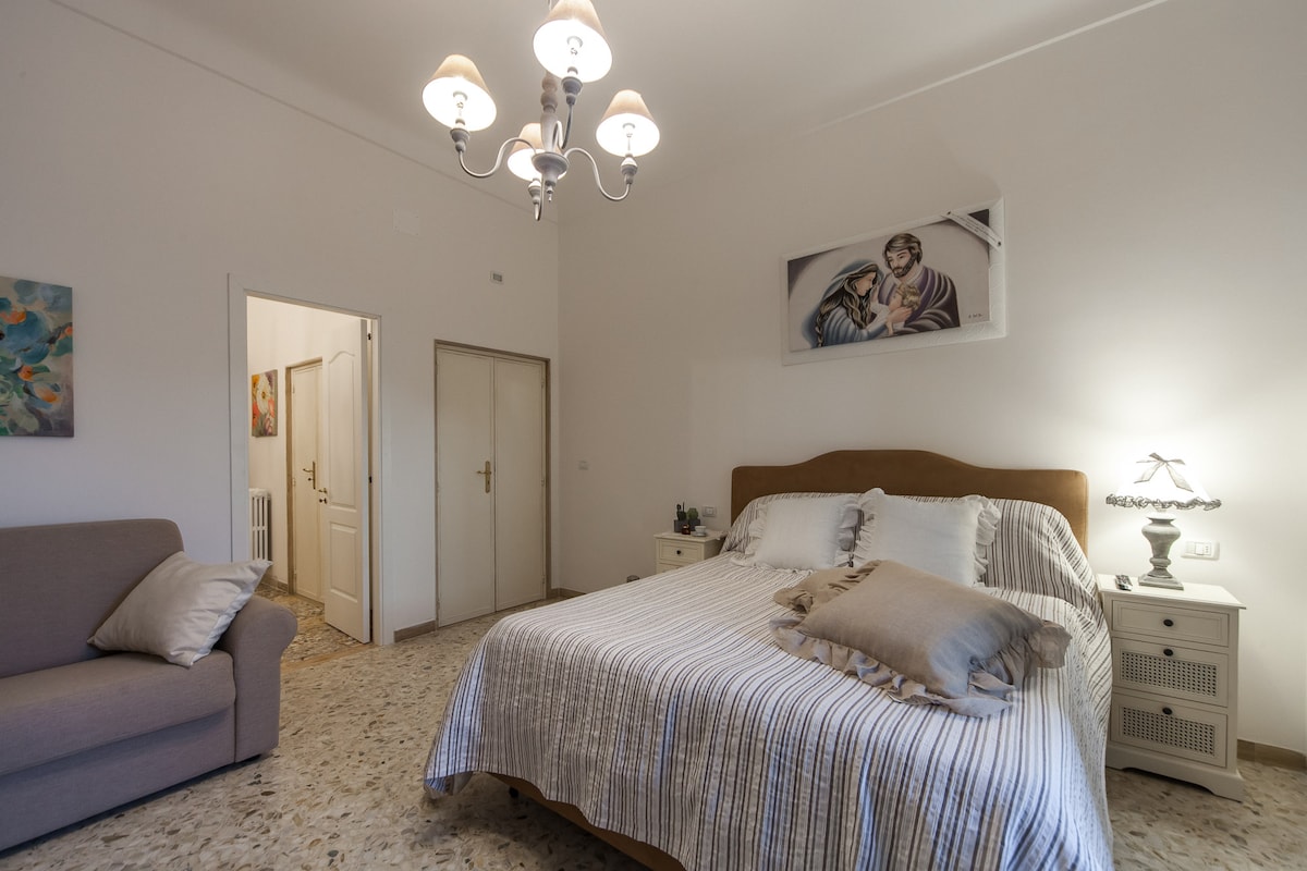 A spacious bedroom features a large bed with a striped coverlet and a decorative pillow. A soft sofa is positioned beside the bed. Soft lighting is provided by a chandelier and bedside lamp. A framed artwork hangs above the headboard, creating an inviting ambiance.