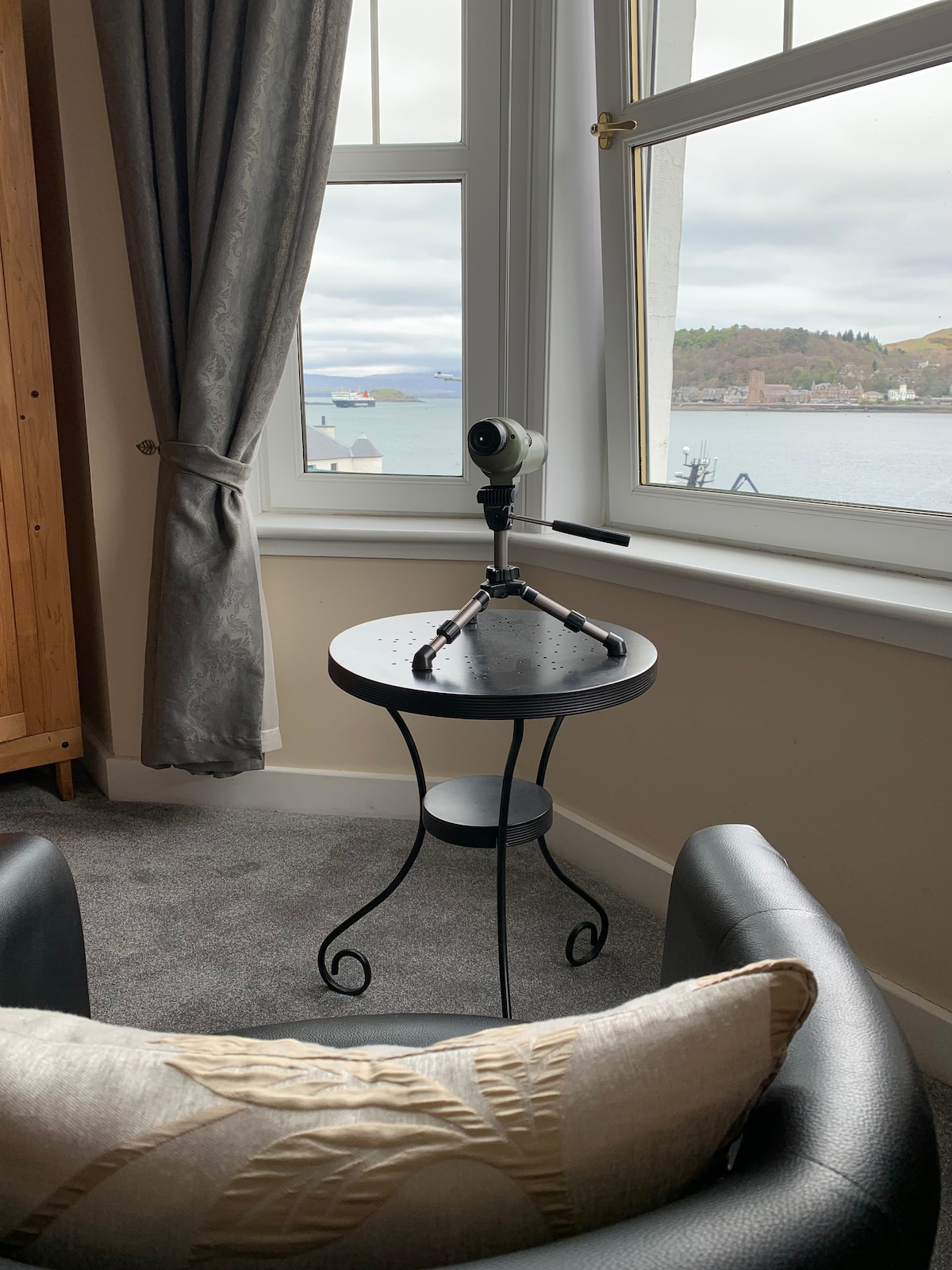 A comfortable seating area is accented by a black round table displaying a telescope, positioned by a large window that offers views of the serene bay. The soft gray curtains frame the window, while a plush chair with a decorative pillow invites relaxation.