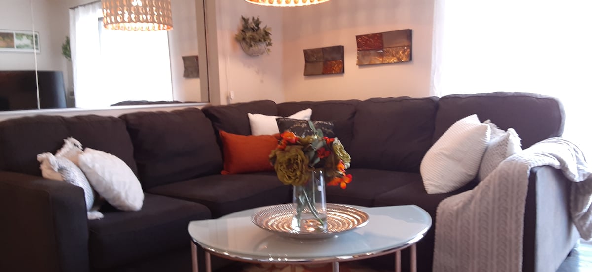 A cozy living area is presented with a sectional sofa adorned with a mix of textured and patterned cushions. A glass coffee table sits at the center, featuring a decorative vase filled with colorful flowers. Soft lighting is provided by an elegant chandelier overhead.