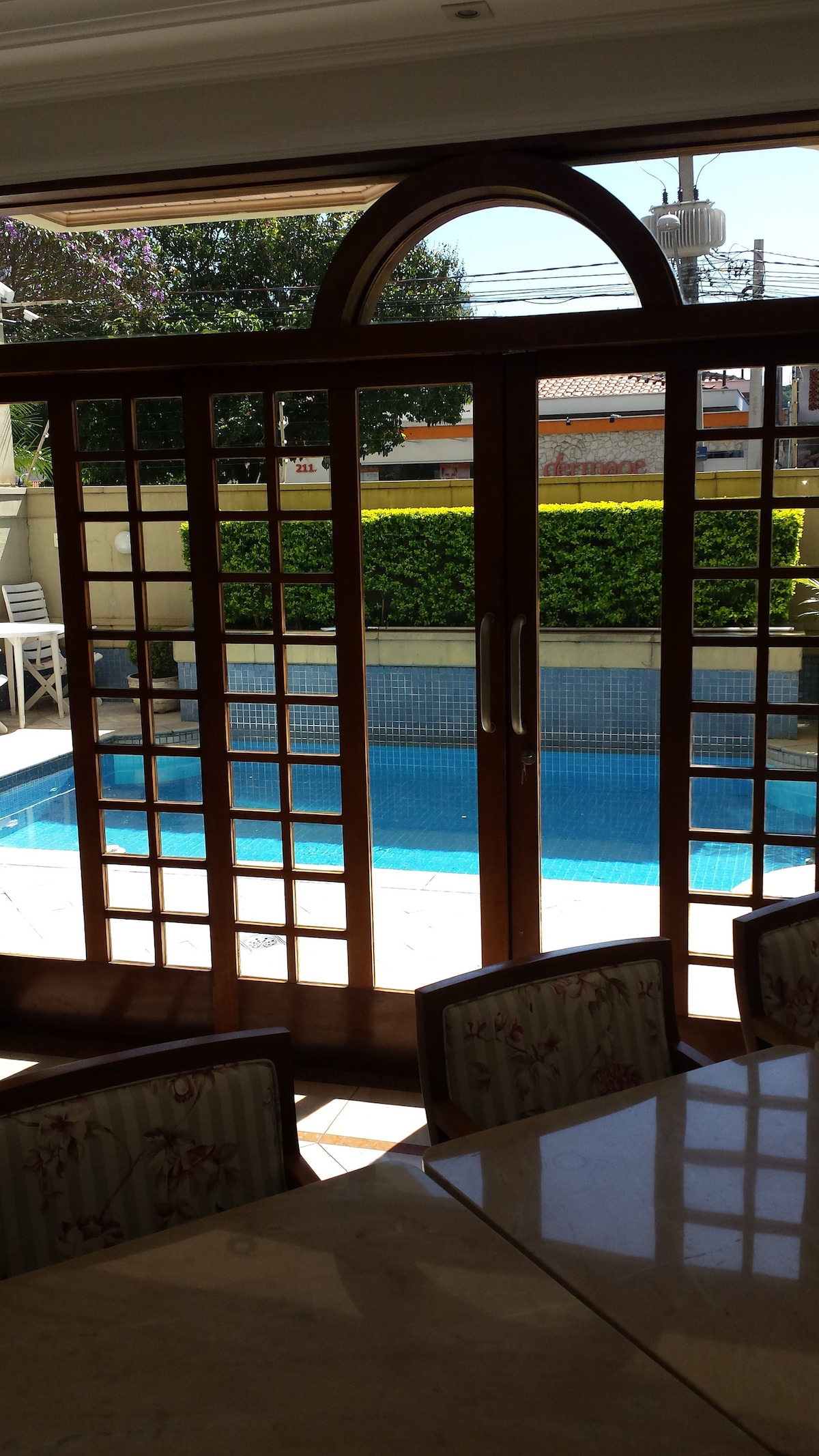 A set of wooden double doors with a grid design opens to reveal a view of a swimming pool surrounded by greenery. The pool reflects sunlight, creating a serene atmosphere. The interior space features a dining table with elegantly patterned chairs.