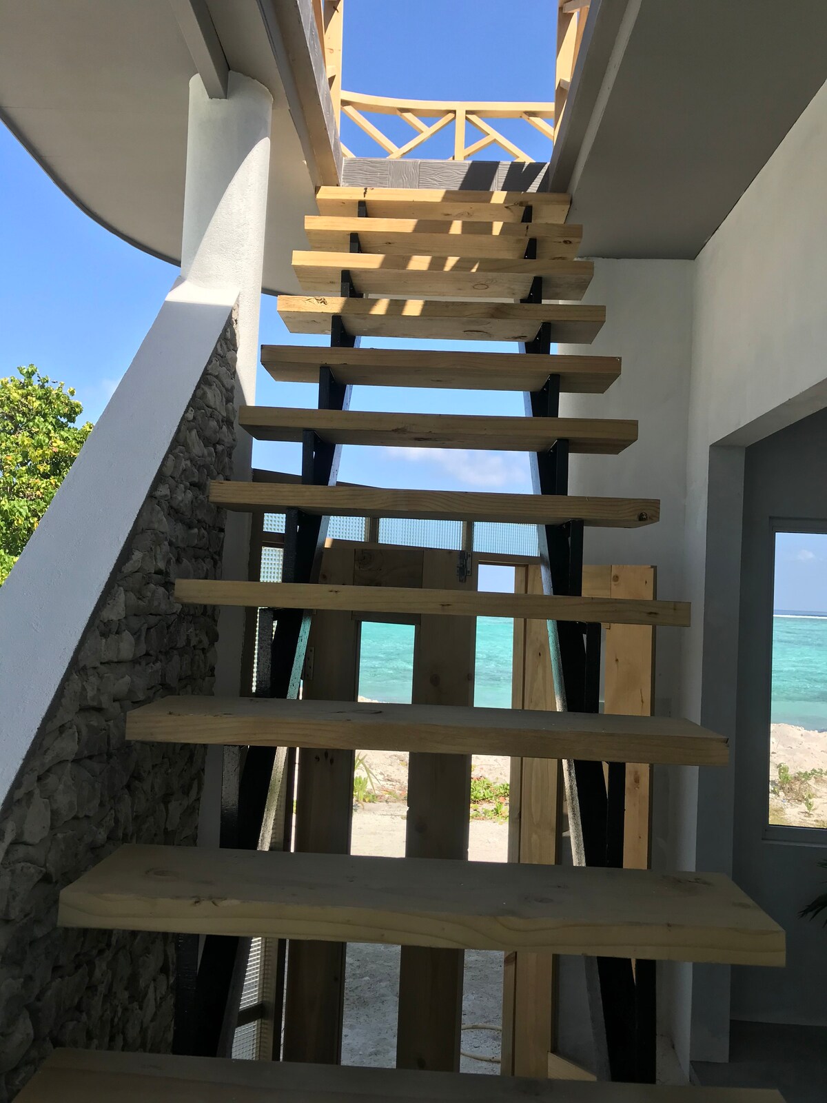 A wooden staircase with a modern design leads towards a bright open space, framed by large windows that overlook the ocean. Natural light floods the area, highlighting the sleek lines and materials used in the construction.