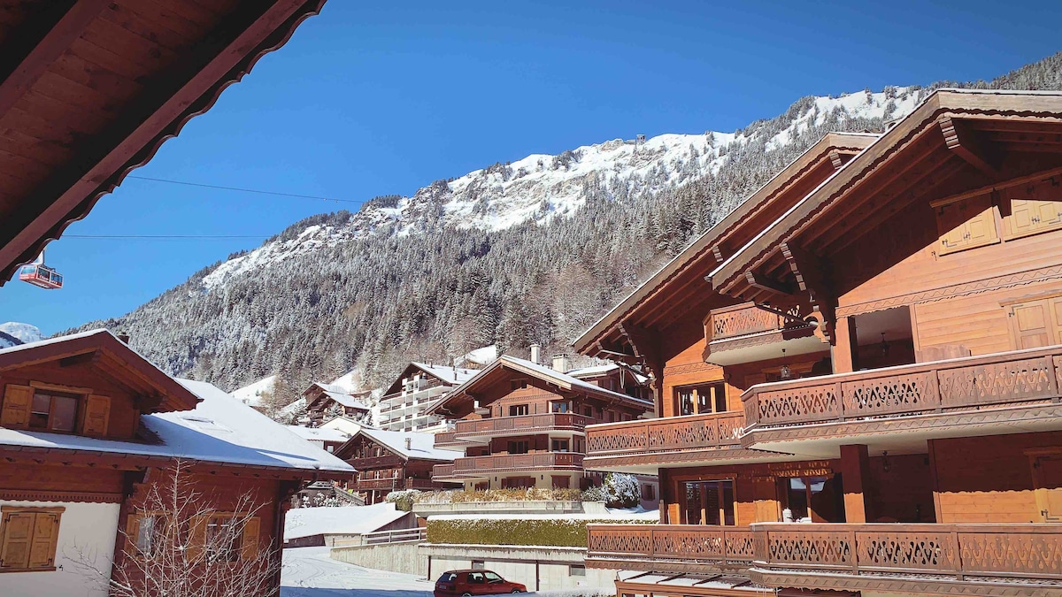 A scenic view captures traditional wooden chalets surrounded by snow-covered mountains. The clear blue sky enhances the serene atmosphere, while the rooflines of the chalets showcase intricate wooden details and balconies. Nearby cable cars can be seen ascending toward the peaks.