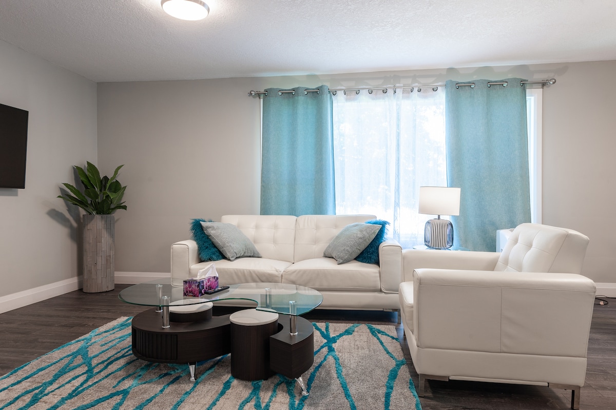 A modern living area features a white sectional sofa accompanied by a matching armchair. A circular glass coffee table is centered on a patterned rug. Light blue curtains frame a large window, allowing natural light to fill the space. A decorative plant adds a touch of greenery.