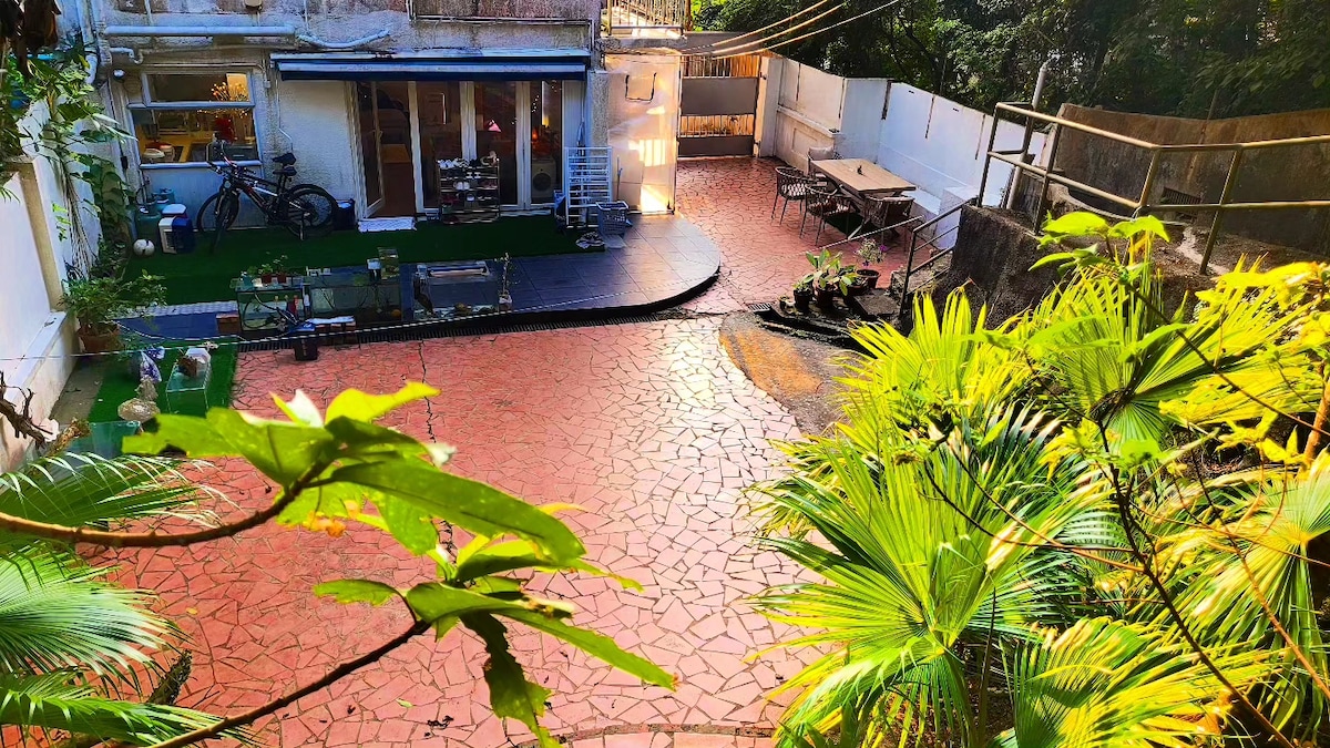A spacious outdoor area features a bright red stone patio surrounded by lush greenery. Tables and chairs are arranged in a friendly setting, while a few small plants are neatly positioned. The area is well-lit with sun illuminating the space, adding a vibrant touch.